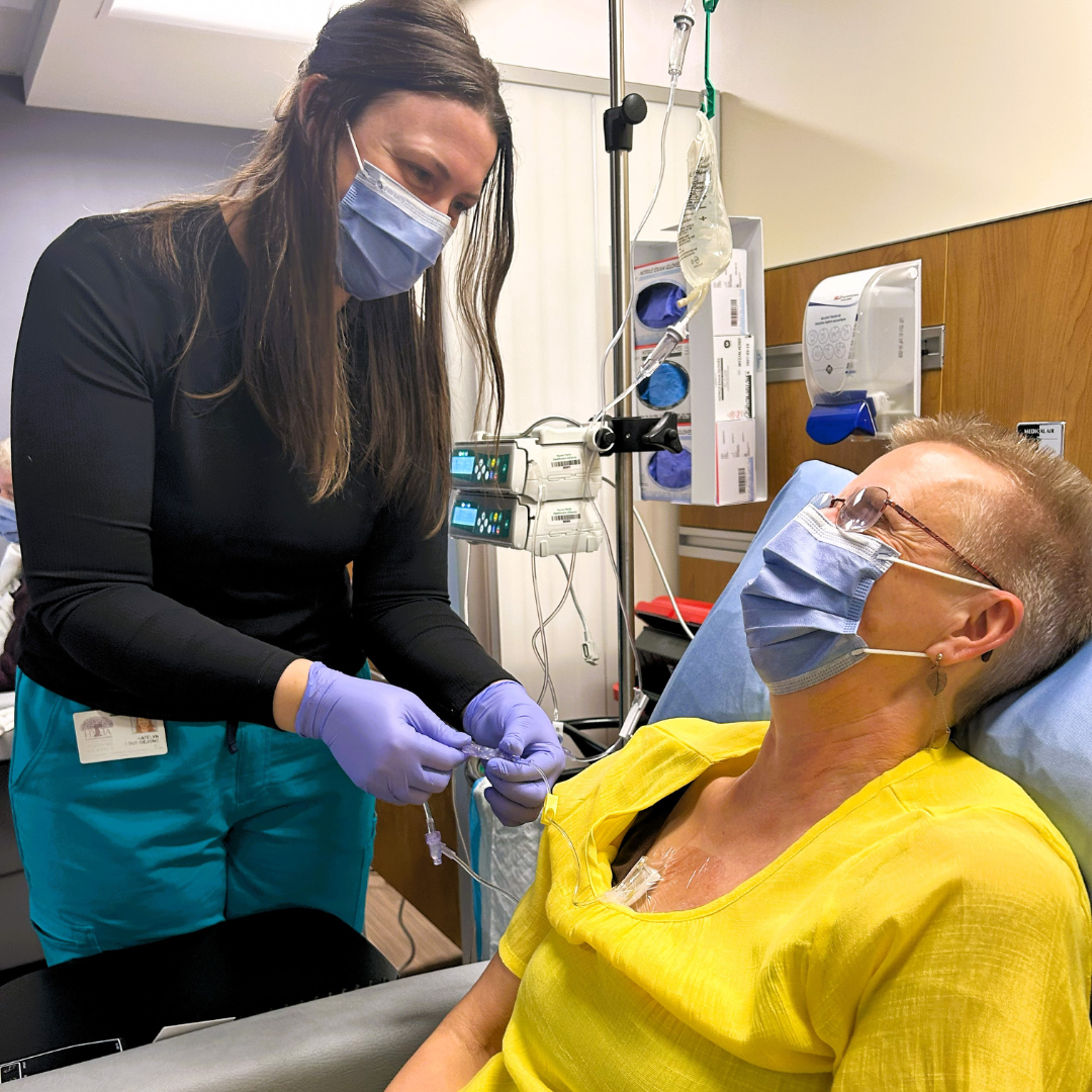 A masked nurse prepares to administer treatment to a patient seated in a reclining chair in the new Cancer and Medical Care Clinic. The patient, also masked and wearing a bright yellow top, looks up at the staff member with a calm expression. IV equipment and medical supplies are visible in the background, highlighting the clinical care setting.